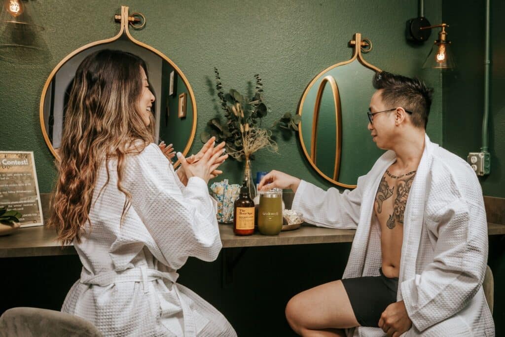 A couple applies lotion while wearing Oakwell bathrobes and enjoying craft beverages