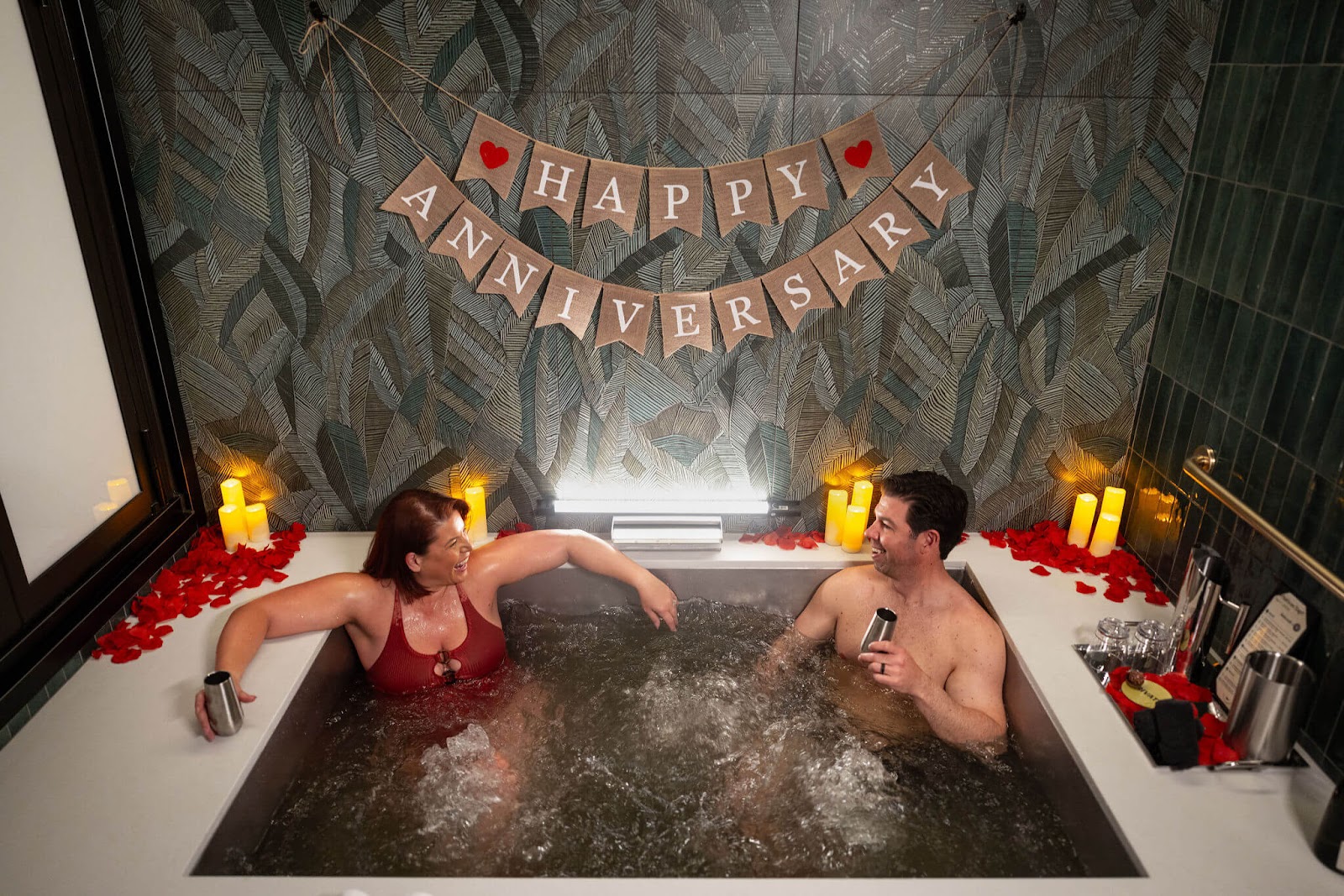 A couple relaxing in Oakwell Beer Spa's beer bath surrounded by candles, rose petals, and a happy anniversary sign