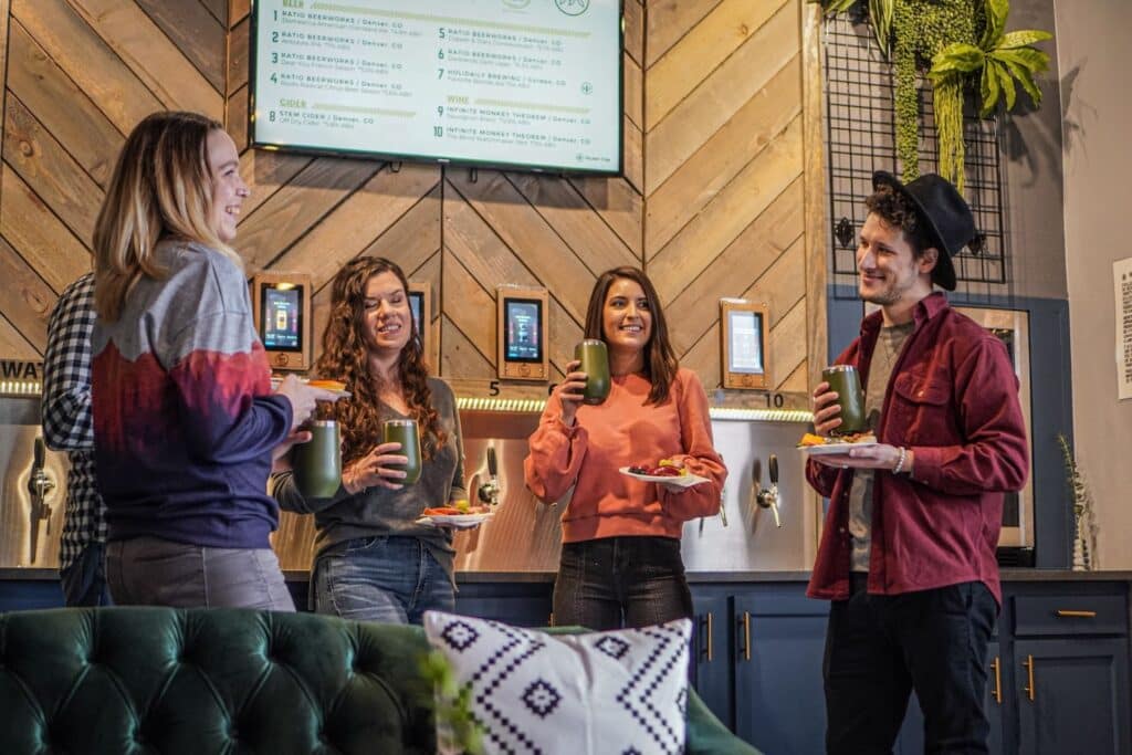 A group of friends gathers around the Oakwell beer wall with beers and snacks.